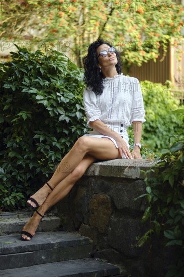 A woman sits elegantly on a stone wall in a vibrant garden, wearing a chic outfit and sunglasses. Sunlight filters through the leaves, creating a serene and relaxing atmosphere