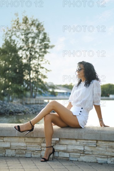A woman with curly hair relaxes on a stone bench by the water, dressed in a chic white outfit and stylish black heels. The scene captures a leisurely moment under a bright sky, surrounded by greenery