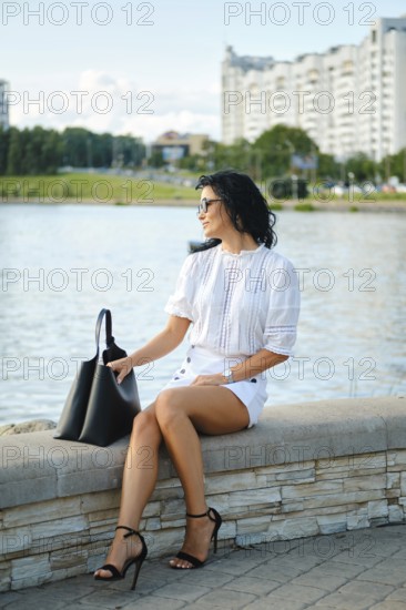 A fashionable woman sits by the water, dressed in a white top and shorts while wearing stylish heels. She gazes towards the cityscape, exuding confidence against a sunny backdrop