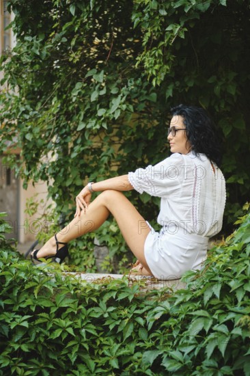 A woman with dark hair sits peacefully in an urban garden surrounded by vibrant green leaves. She wears a white outfit and sunglasses, exuding a relaxed vibe on a sunny day