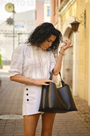 A woman with curly hair stands in a charming alleyway, wearing a white blouse and a stylish skirt. She is looking for something into her black handbag