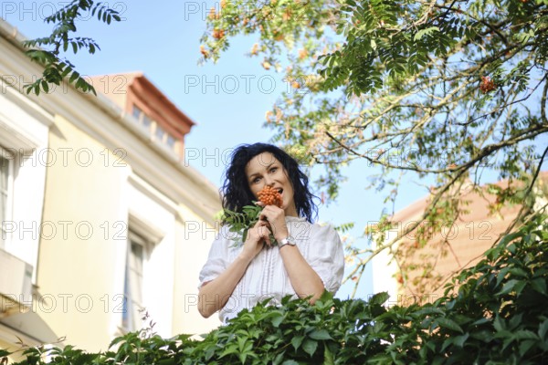 A playful woman stands surrounded by greenery holding a cluster of bright berries. The sun shines down, highlighting her joyful expression as she enjoys the beauty of the garden setting