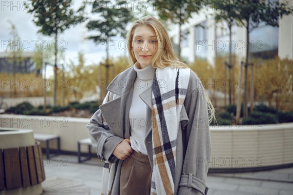 A young woman stands outdoors, showing off her fashionable gray coat featuring a plaid pattern. She exudes confidence while dressed in a chic outfit amidst a well-maintained urban landscape