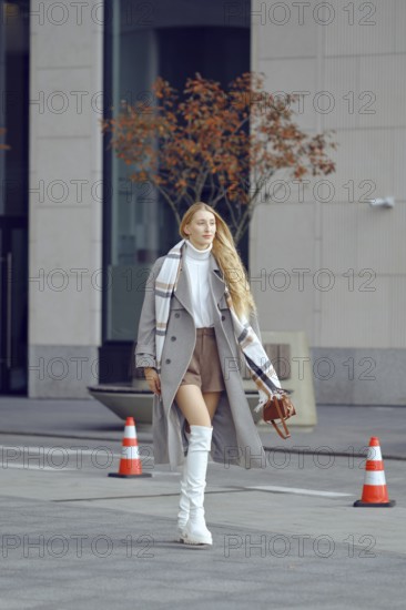 A tall model walks down a modern city street, showcasing chic winter fashion with a stylish overcoat, knee-high boots, and a patterned scarf
