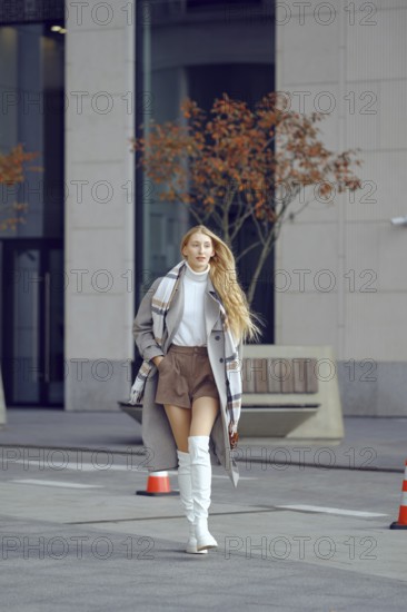 A confident woman strolls along a city street, wearing a trendy autumn outfit that includes boots and a chic coat