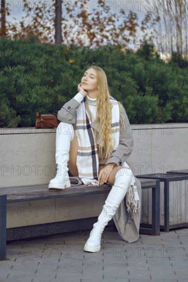 A dreamy woman sits thoughtfully on a bench in a park, dressed in a fashionable outfit with a long coat and scarf. She enjoys the serene environment, surrounded by greenery and a pleasant atmosphere