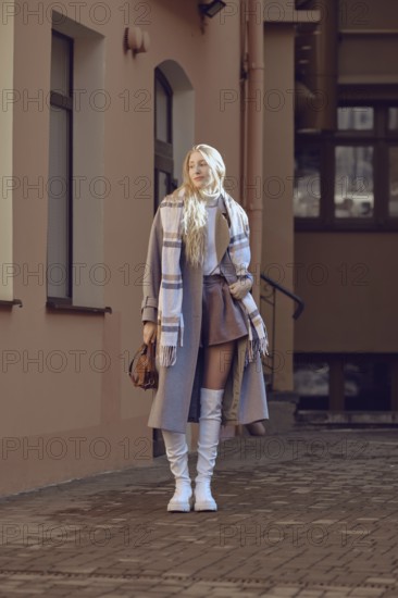 A fashionable young woman in wool coat, knee-high boots and scarf posing in a sunny urban alley. Her casual elegance showcases a perfect blend of comfort and style