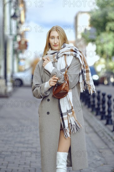 A young woman with blonde hair stands on a cobblestone street, wearing a fashionable coat and a scarf wrapped around her neck. She appears relaxed and confident, enjoying an autumn day