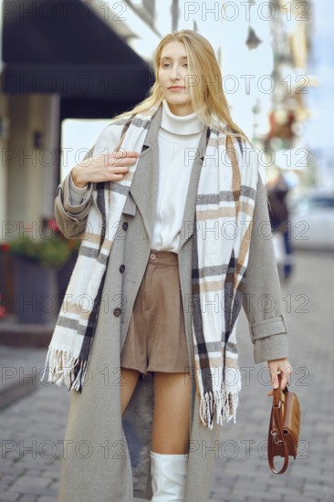 A fashionable young woman strolls down a cobblestone street, wearing a long coat and a plaid scarf, complemented by shorts and knee-high boots. The urban setting is lively with a sunny ambiance
