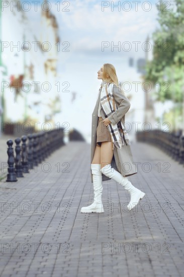 Stylish woman in a long coat and knee-high boots strolls confidently cross a charming cobblestone street. The vibrant surroundings highlight her chic style