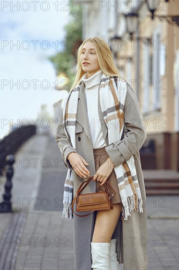 City portrait of a fashionable confident woman on a picturesque street lined with vintage architecture, dressed in a chic coat and a striped scarf. Her stylish handbag complements her outfit