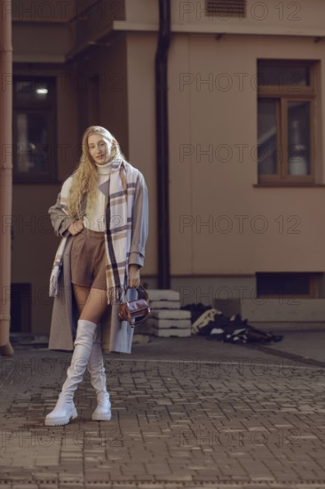A fashionable woman stands confidently on a cobblestone street, wearing knee-high boots and a stylish coat with a scarf
