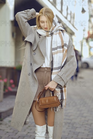 Stylish woman in beige coat and white boots walking down the street stopped to adjust her scarf