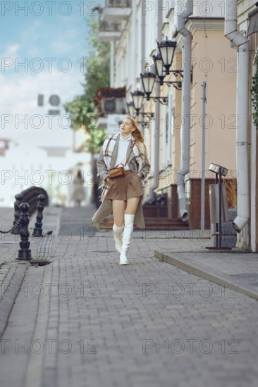 A young woman walks confidently down a cobblestone street wearing a stylish trench coat and knee-high boots