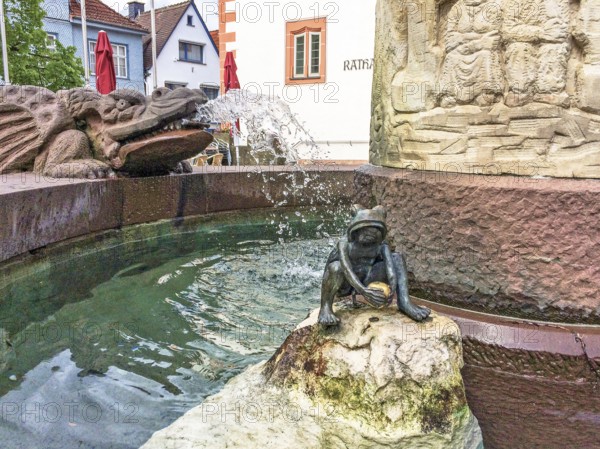 Sculpture figure of a frog holding a golden ball depicting the Frog King from the Brothers Grimm fairy tale in a fairy tale fountain, gargoyle in the form of a winged dragon behind it on the left, Steinau an der Straße, Hesse, Germany