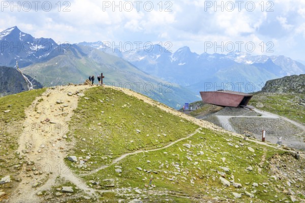 View of left hills Hiking trails lead to the summit with Christian cross, centre right Timmelsjoch pass museum made of reddish-coloured concrete at the top of the mountain pass Alpine pass Alpine mountain road Alpine road Pass road Border pass Pass Timmelsjoch Passo del Rombo in Alps, Timmelsjoch High Alpine Road, Province of Tyrol, Austria, South Tyrol, Italy