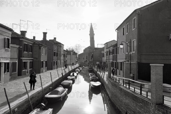 Strollers along the canal, typical street scene in winter, monochrome, facades in Burano, leaning campanile of the church of San Martino, island in the lagoon of Venice, Italy