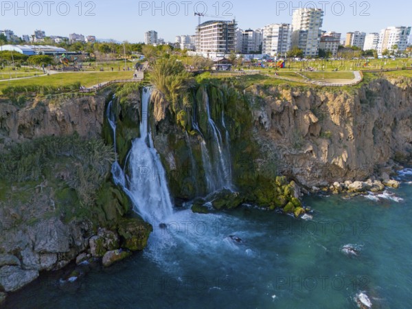 A waterfall cascades from a cliff into the sea, surrounded by urban landscape and green parks, aerial view, Lower Düden Waterfall, Düden River, Antalya, Turkey
