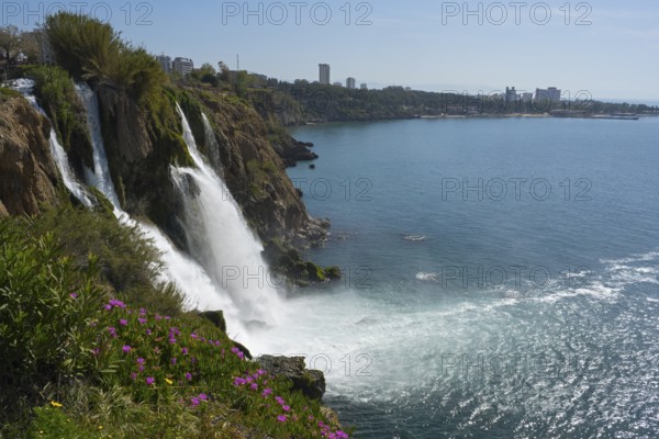 A waterfall cascades from cliffs into the sea, surrounded by flowers and a town in the distance, Lower Düden Waterfall, Düden River, Antalya, Turkey