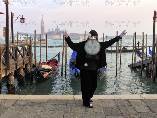 Person with black costume and mask, full moon mask, boat dock at St Mark's Square, behind the church of San Giorgio Maggiore, Carnival in Venice, Italy