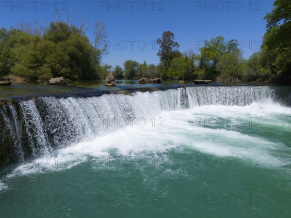 A waterfall flows into a turquoise-coloured river, surrounded by green vegetation and blue sky, aerial view, Manavgat Waterfall, Manavgat River, Side Turkey