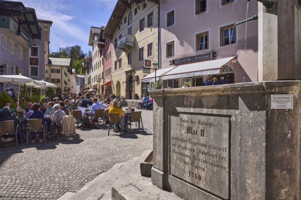 Market fountain, stone fountain, pedestrian zone, stairs, general architecture, historical residential and commercial buildings, outdoor area of a restaurant, pedestrians and guests as secondary motif, hill, trees, blue sky, cirrostratus clouds, market square, Berchtesgaden, Upper Bavaria, district Berchtesgadener Land, Bavaria, Germany