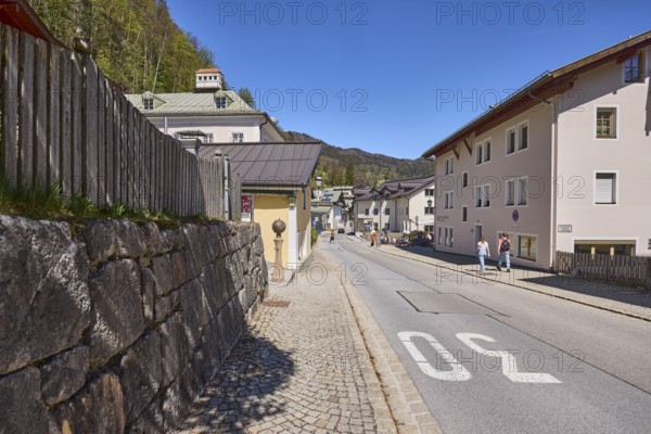 General architecture, apartment buildings, residential building, pavement, cobblestones, street, zone 30 marking, quarry stone wall, wooden fence, mountain landscape, hill, forest, pedestrian as secondary motif, blue sky, cloudless, Nonntal, Berchtesgaden, Upper Bavaria, district Berchtesgadener Land, Bavaria, Germany