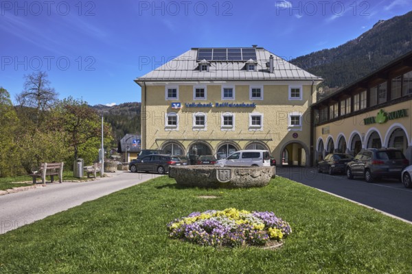 Volksbank Raiffeisenbank Oberbayern Südost eG, Fischerbrunnen, master stonemason August Wolf, fountain, building, car park, vehicles, lawn, flower bed, trees, mountain landscape, mountains, coniferous forest, blue sky, cirrostratus clouds, Am Fischerbichl, Bahnhofstraße, Berchtesgaden, Upper Bavaria, Berchtesgadener Land district, Bavaria, Germany