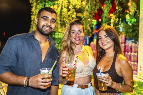 Three friends smiling and holding cocktails at a vibrant outdoor night party, enjoying the festive atmosphere