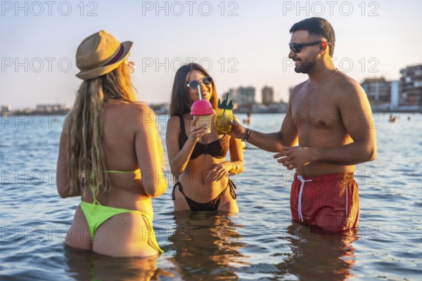 Three friends enjoying refreshing cocktails while standing in ocean water at sunset, celebrating summer vacation together