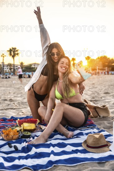 Two women enjoying a beach picnic at sunset, capturing a joyful selfie surrounded by fresh fruit, snacks, and laughter