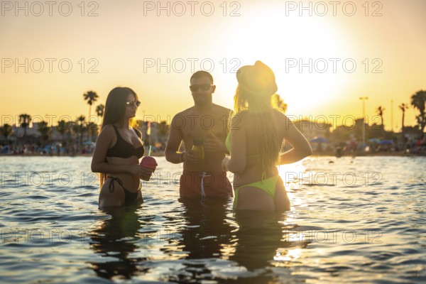 Three friends are having cocktails in the ocean at a beach party, enjoying the beautiful sunset