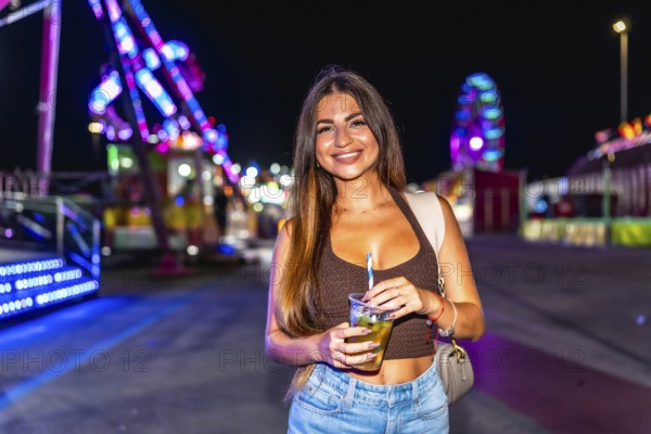 Young woman holding a refreshing drink at a lively amusement park, enjoying the vibrant atmosphere and colorful lights