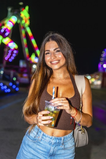 Young woman holding a refreshing cocktail at an amusement park, enjoying the vibrant night atmosphere
