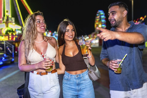 Group of friends enjoying cocktails, walking and laughing together at an amusement park, surrounded by colorful lights and festive energy