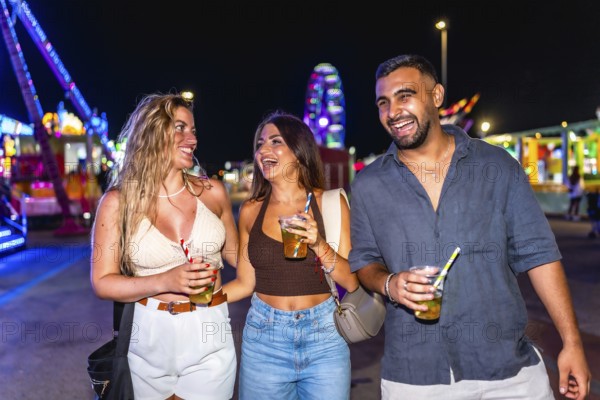 Three friends walking and laughing together at amusement park at night, holding drinks and enjoying their time