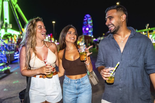 Three joyful friends walking and laughing together at an amusement park at night, enjoying colorful cocktails under vibrant lights