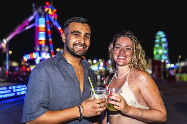 Young man and woman smiling, holding cocktails, enjoying a night out at amusement park with colorful lights in background