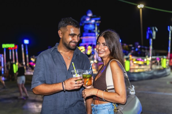 Young couple smiling and holding colorful cocktails at a lively funfair, celebrating nightlife and enjoying joyful moments together