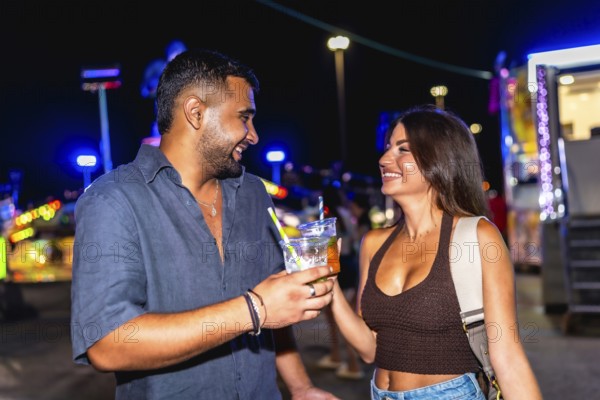Young man and woman smiling and toasting with colorful cocktails while enjoying the vibrant atmosphere of an amusement park at night