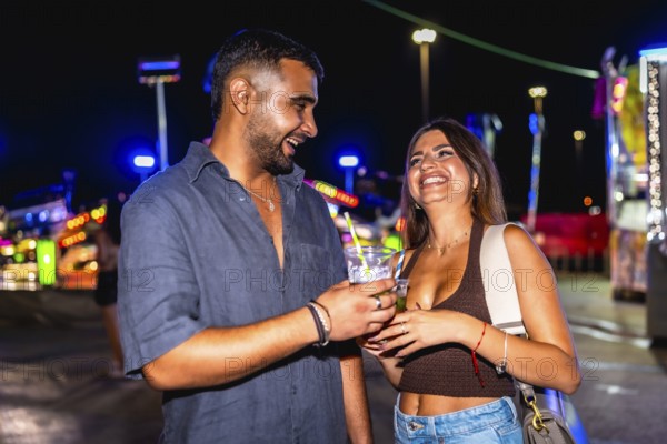 Young man and woman laughing joyfully while enjoying cocktails together, surrounded by the vibrant lights of an amusement park at night