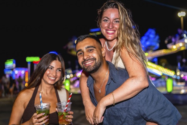 Three friends are having fun at an amusement park at night, enjoying cocktails and the festive atmosphere