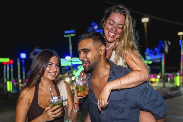 Group of cheerful friends enjoying cocktails and laughing together at an amusement park during a vibrant summer night filled with excitement