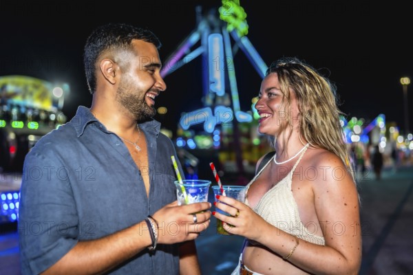 Happy couple enjoying drinks at amusement park, celebrating summer night with colorful lights and fun attractions in the background