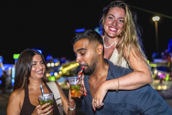Three friends are having cocktails at an amusement park at night, enjoying the vibrant atmosphere and colorful lights