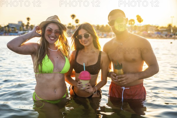 Three friends enjoying refreshing cocktails while standing in ocean water during a vibrant sunset on a tropical beach