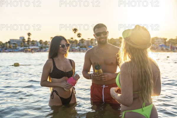 Three friends are having refreshing cocktails in the ocean during a beautiful sunset at the beach
