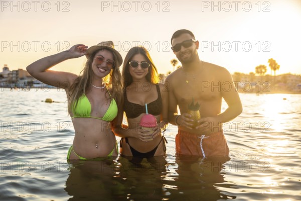 Three friends are having fun in the ocean at sunset, enjoying refreshing cocktails and the golden hour light