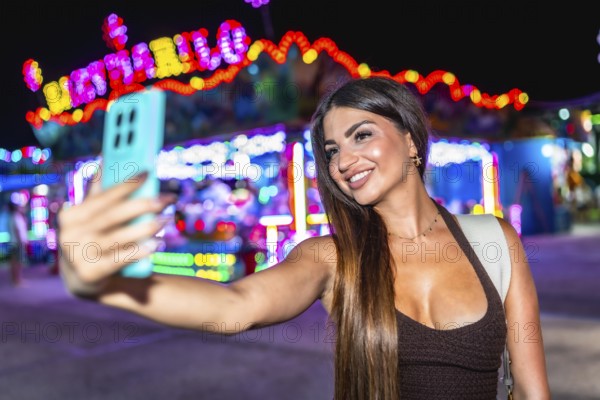 Happy woman enjoying a vibrant night at an amusement park, taking a selfie with her smartphone amidst colorful lights and festive atmosphere