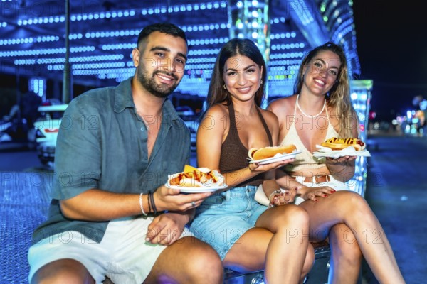 Three friends are sitting and smiling, holding hot dogs on paper plates at a brightly lit amusement park at night